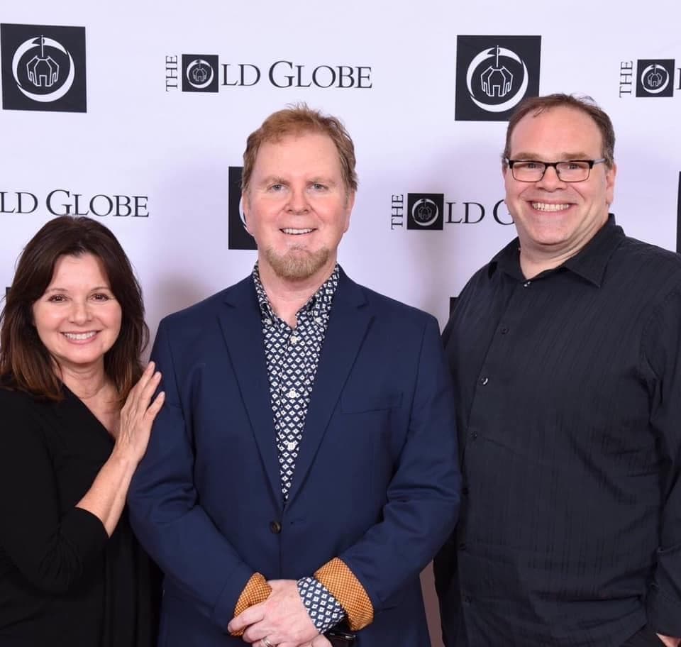 Three people smile in front of a backdrop printed with The Old Globe's logo.