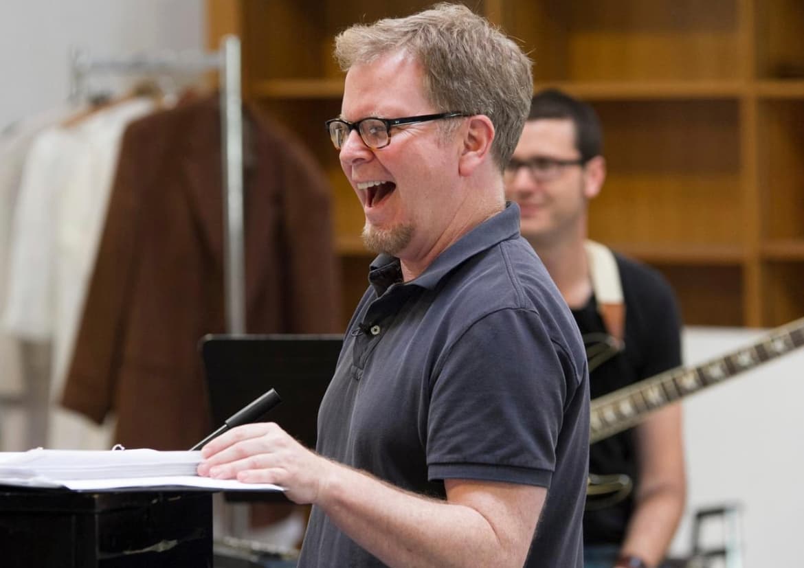 A man laughs at a music stand during rehearsal while a guitarist plays behind him.